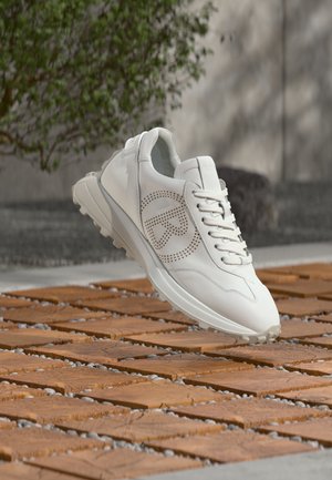 Zapatilla deportiva blanca con logo perforado en forma de "R", cordones y suela texturizada, suspendida sobre una superficie de madera con patrón al aire libre.