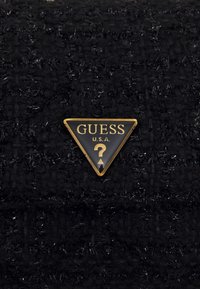 Zwarte textuurrijke stof met een driehoekig gouden logo met de tekst "GUESS" en "U.S.A." gedrukt, geaccentueerd door een centraal vraagtekenontwerp.