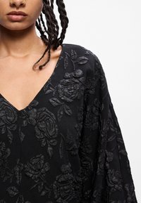 Melna auduma blūze ar V-veida kaklu, reljefa rozā rakstu, plūstošām piedurknēm un smalkiem ziedu akcentiem. Tekstūra piešķir dziļumu.