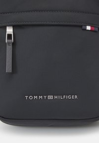 Schwarzer Stoffrucksack mit einem silbernen Reißverschluss, strukturiertem Tragegurt und einem kleinen, mehrfarbigen Logoakzent. Markenname am Boden geprägt.