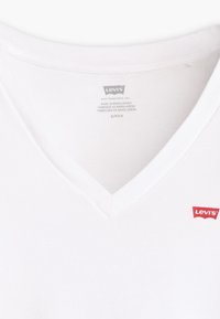 Bijela pamučna majica s V-izrezom, s malim crvenim Levi's logotipom na lijevoj strani prsnog područja. Glatka tekstura, ležeran stil.