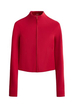 Vrouw staand, draagt een nauwsluitend rood shirt met lange mouwen en legging, met witte sneakers, tegen een effen grijze achtergrond.