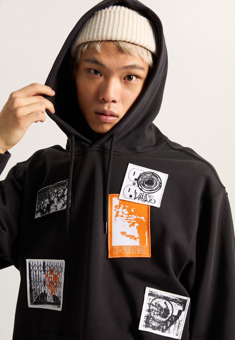 Czarny sweatshirt z kapturem z wieloma naszywkami w białym, szarym i pomarańczowym kolorze, prezentującymi różne grafiki i napisy, w tym "HI-VOLTAGE".