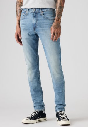 Hombre con vaqueros ajustados de color azul claro y zapatillas negras de caña alta, de pie sobre un fondo liso, manos relajadas a los lados con tatuajes visibles.