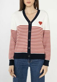 Cardigan rayé rouge et blanc avec un décolleté en V bleu marine et des boutons. Présente un petit emblème en forme de cœur rouge sur la poitrine gauche.