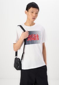 Weißes Baumwoll-T-Shirt mit einem Grafikdesign in roten und marineblauen Streifen, auf dem "JACK & JONES" steht, kombiniert mit einer kleinen schwarzen Umhängetasche.
