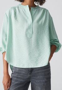 Blusa verde menta chiaro con scollatura tonda, pattina con tre bottoni e spalle scese; presenta un tessuto testurizzato e una vestibilità ampia.