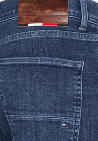 Tmavě modré džíny z denimu s koženou nášivkou označenou "HILFIGER" na pásku kalhot. Vyznačují se klasickým prošitím a detailem na zadní kapse.