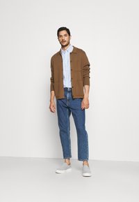 Cardigan marrone sopra una camicia button-up azzurra, abbinato a jeans blu e scarpe slip-on grigie. Design semplice con una vestibilità rilassata.