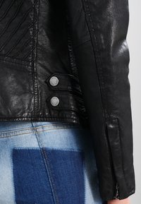 Černá kožená motocyklová bunda s vyšívanou texturou, zipovými manžetami a stříbrnými knoflíky na druk. V kombinaci s džíny s velkou modrou nášivkou.