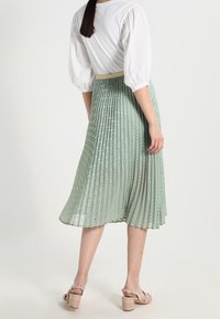 Gonna plissé verde chiaro con pois dorati, di lunghezza al ginocchio, indossata con una maglietta bianca e tacchi beige sottili, con una vita ben definita.