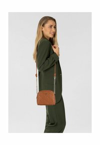 Braune Umhängetasche mit geprägtem Muster, silberner Kettenriemen und runder Form. Von einem Model in einem grünen Anzug über der Schulter getragen.