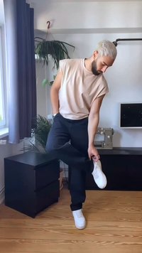 Top senza maniche beige chiaro, pantaloni neri e sneakers bianche. In piedi con un piede su un mobile a cassetti nero, mentre aggiusta la sneaker.