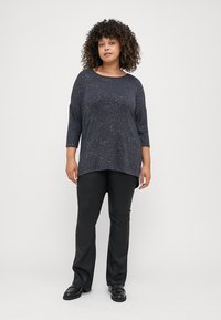 Tengerészkék, hosszú ujjú felső csillogó mintával, fekete nadrággal és loaferekkel párosítva. A felső kerek nyakú és oldalsó hasítékokkal rendelkezik.