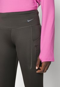 Leggings gris oscuro con cintura alta, que cuentan con un pequeño bolsillo con cremallera, combinados con una camiseta de manga larga rosa. Textura suave. Logo de Nike presente.
