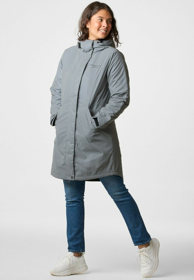 Grijze lange jas met ritssluiting en drukknoopsluiting, capuchon en contrasterende zwarte accenten, gecombineerd met blauwe jeans en witte sneakers.