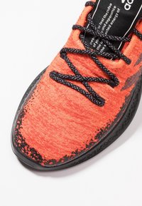 Oranžová sportovní obuv s texturovaným pleteným svrškem, černými akcenty a kulatými tkaničkami. Má hladkou černou gumovou podrážku s jemným vzorem.