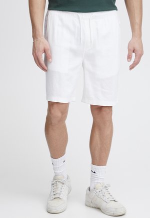 Uomo che indossa pantaloncini bianchi con coulisse, calzini bianchi alti, scarpe da ginnastica bianche e una maglietta verde scuro, in piedi contro uno sfondo semplice.