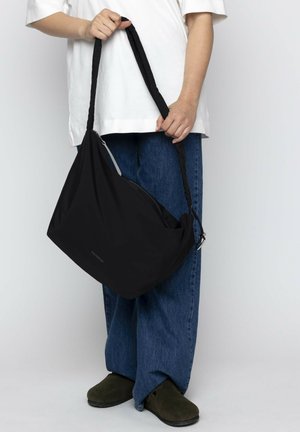 Bolso de mensajero negro con una textura suave, tirante único y detalle de cremallera plateada, sostenido en la mano, combinado con denim y zuecos.