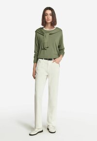 Zielony sweter z opadającym kołnierzem i długimi rękawami, zestawiony z białymi spodniami o prostym kroju i białymi mokasynami. Proste, teksturowane materiały.