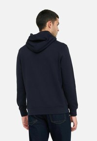 Homme portant un sweat à capuche noir uni et un jean bleu foncé, tourné vers le fond gris clair.