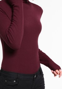 Burgundské žebrované rolákové tričko s dlouhými rukávy, slim fit a otvory na palce. V kombinaci s černými kalhotami, které mají hladkou strukturu.