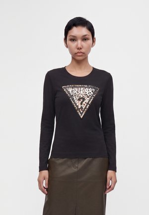 Femme aux cheveux courts et foncés portant un haut noir à manches longues avec un logo Guess imprimé léopard et une jupe en cuir vert olive, debout devant un fond blanc.