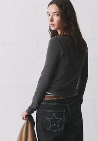 Junge Frau mit langen dunklen Haaren, die einen dunklen Langarmoberteil und schwarz gestickte Jeans trägt, hält einen beigen Mantel vor einer weißen Wand.