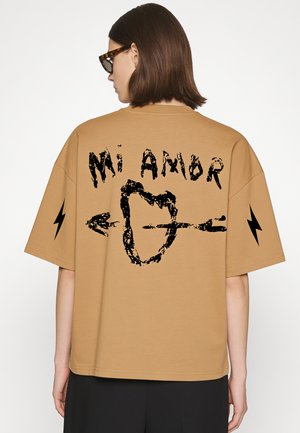 Person with straight brown hair and tortoiseshell glasses wearing tan shirt with black text "MI AMOR" and heart pierced by arrow, black lightning bolts on sleeves.
