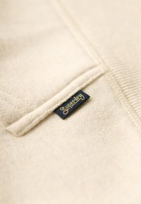 Tessuto del felpa beige con una superficie leggermente strutturata. Presenta un'etichetta "Superdry" cucita di colore blu scuro e oro sul lato.