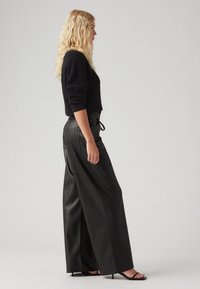 Črn rebrast pulover v kombinaciji z visokimi črnimi usnjeni širokimi hlačami. Model nosi črne sandale s trakovi, kar poudari njen eleganten profil.