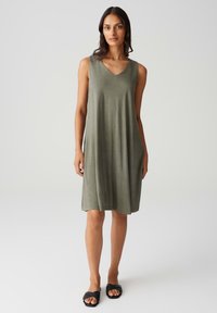 Sleeveless olive green dress with a V-neck design, made of soft fabric, falls to knee length. Worn with black flat sandals.