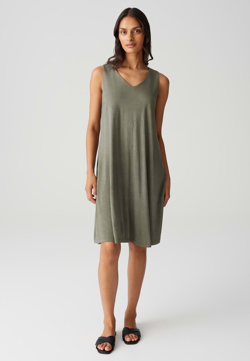 Sleeveless olive green dress with a V-neck design, made of soft fabric, falls to knee length. Worn with black flat sandals.