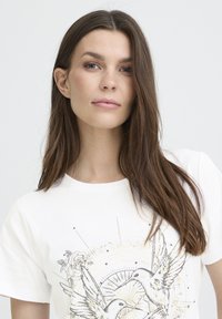 White cotton t-shirt featuring a detailed print of two hummingbirds surrounded by floral and geometric gold accents. Short sleeves, round neckline.