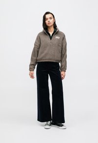 Fleecový pulóver v taupe s polovičným zipsom a logom "GAP", kombinovaný so širokými čiernymi manšestrovými nohavicami a klasickými čierno-bielymi teniskami.