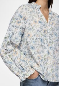 Blouse florale en bleu clair avec des accents jaunes. Présente un devant à bouton, un col froncé et des manches longues resserrées. Matériau en coton.