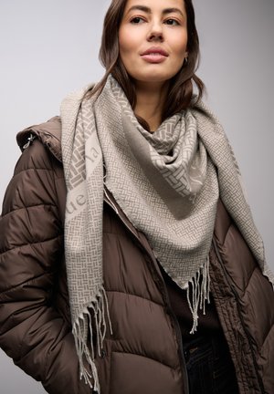 Écharpe beige à motifs avec bords à franges, enroulée autour du cou, assortie à une doudoune marron. Détail du tissu texturé visible.