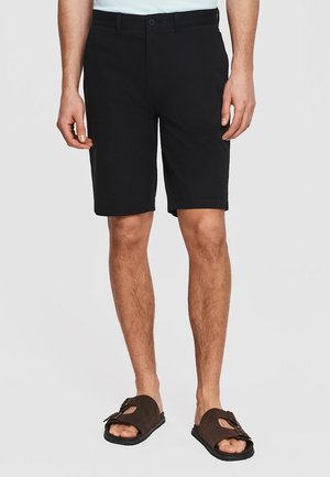 Mężczyzna w czarnych, sięgających do kolan szortach i ciemnobrązowych sandałach z odkrytymi palcami, stojący na jednolitym jasnym tle.