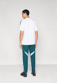 White athletic t-shirt with black stripes on shoulders, paired with teal pants featuring white accents and side zippers, worn with black shoes.