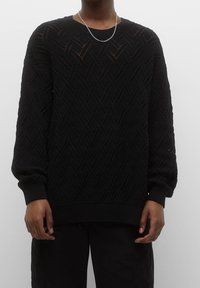 Zwarte gebreide trui met een textuur van een diamantpatroon, brede geribde manchetten en een ronde hals. Draagt zwarte broek en een zilveren ketting.