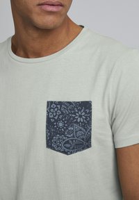 Tricou din bumbac verde deschis cu guler rotund și o buzunară cu model albastru închis, având un design floral și paisley.