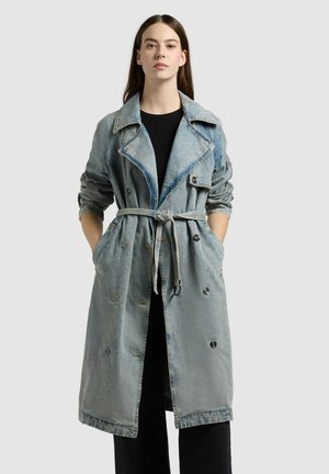 Žena oblečená v svetlomodrom rifľovom trenčkotu s vyhrnutými rukávmi a zviazaným opaskom, stojaca s rukami v kapsách pred jednofarebným sivým pozadím.