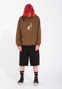 Felpa con cappuccio marrone e stampa grafica, pantaloni corti neri, calze a motivo e scarpe nere, in piedi su uno sfondo bianco.