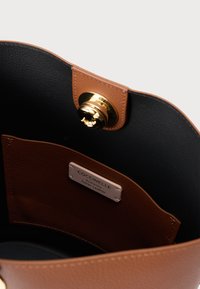 Εσωτερικό καφέ δερμάτινης τσάντας, με λεία μαύρη επένδυση και κούμπωμα σε χρυσό τόνο. Περιλαμβάνει ετικέτα που αναγράφει «Γνήσιο Ιταλικό Δέρμα».