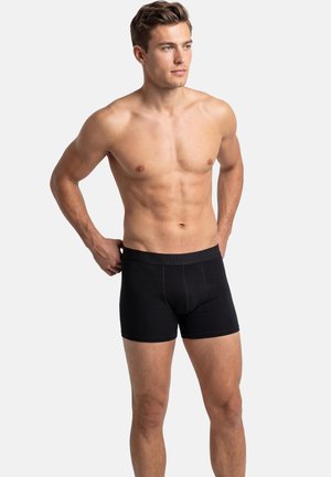 Barbent mand med kort hår iført sorte boxershorts, der står med hænderne på hofterne og kigger let til siden mod en hvid baggrund.