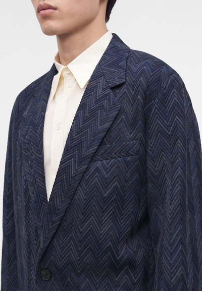 Homme portant un blazer bleu foncé à motif chevron sur une chemise blanche boutonnée, montré du cou à la poitrine sur un fond uni.
