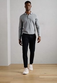 Grijze long-sleeve polo, zwarte cargobroek en witte sneakers. Rechte pasvorm met zachte texturen en subtiele details, lopend op houten vloeren.