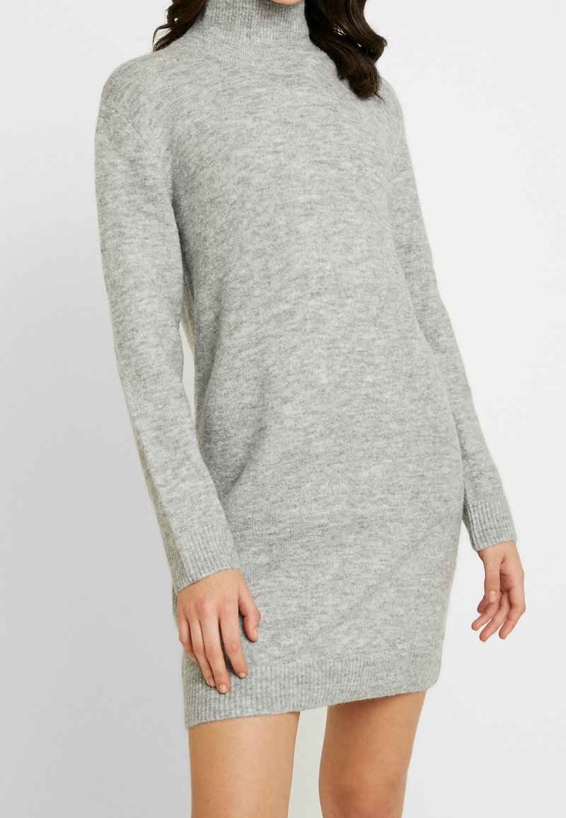 Femme portant une robe pull à col roulé en maille douce grise à manches longues, debout devant un fond clair.