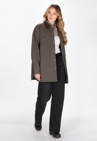 Donna con giacca oversize oliva, sciarpa beige, top bianco, pantaloni larghi neri e scarpe nere, in piedi davanti a uno sfondo bianco.