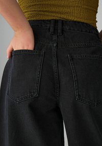 Jeans de mezclilla negra con cintura alta, que presentan dos bolsillos traseros y costuras visibles. La tela tiene una apariencia suave pero texturizada.